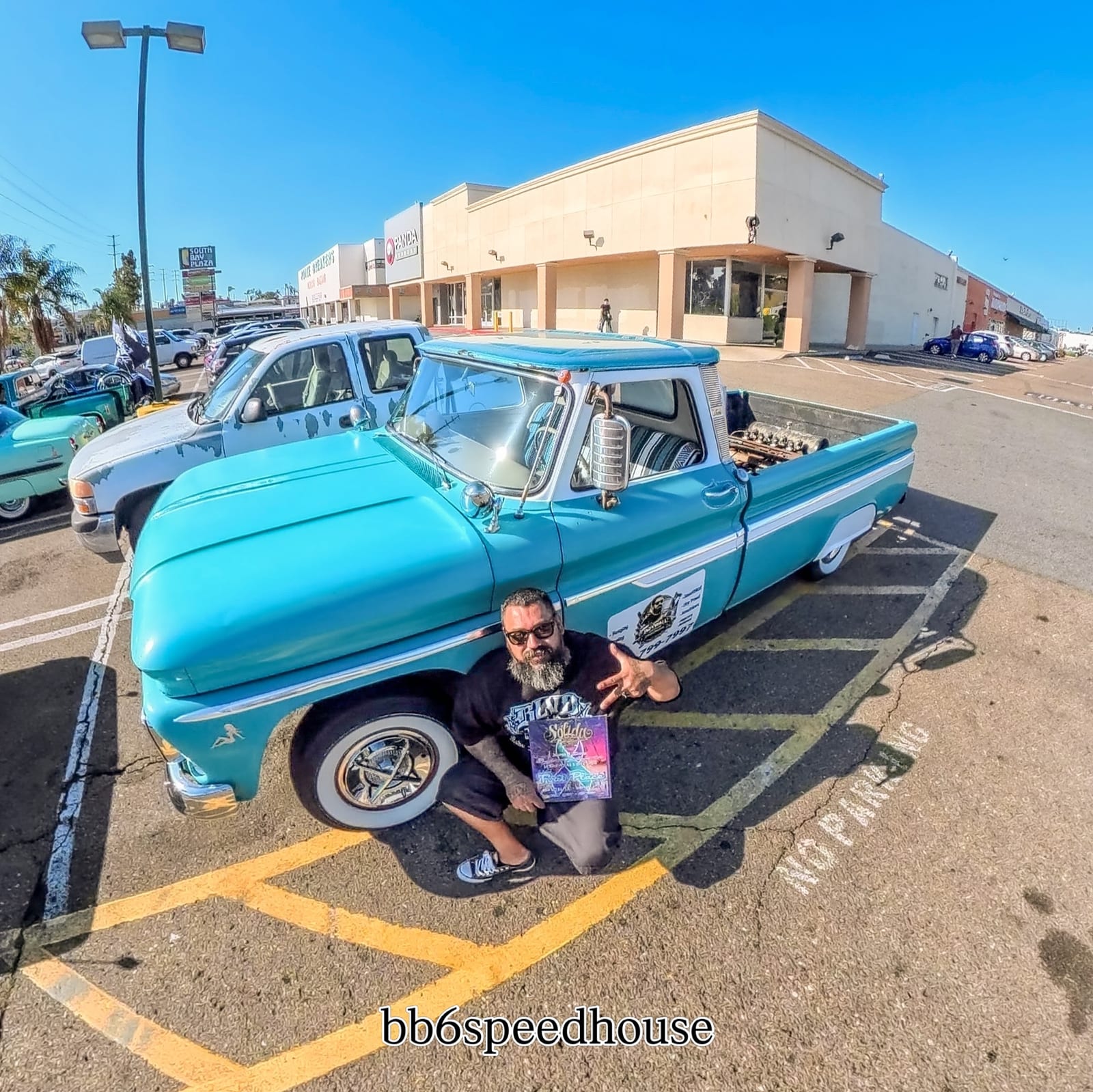 A club supporter poses beside a teal classic pickup at a lowrider gathering.